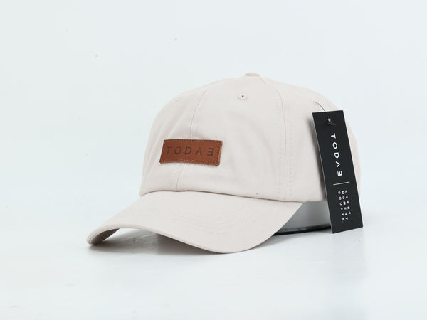 A light beige baseball cap with a brown leather patch on the front displaying the brand name 'TODAE'. A black paper hangtag is attached to the side of the brim.